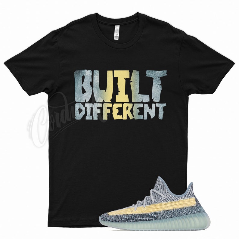 Puede incluir: Una camiseta negra con el texto "BUILT DIFFERENT" en letras amarillas y azules. Un par de zapatillas de deporte azul claro y amarillas se encuentran en la esquina inferior derecha de la imagen.