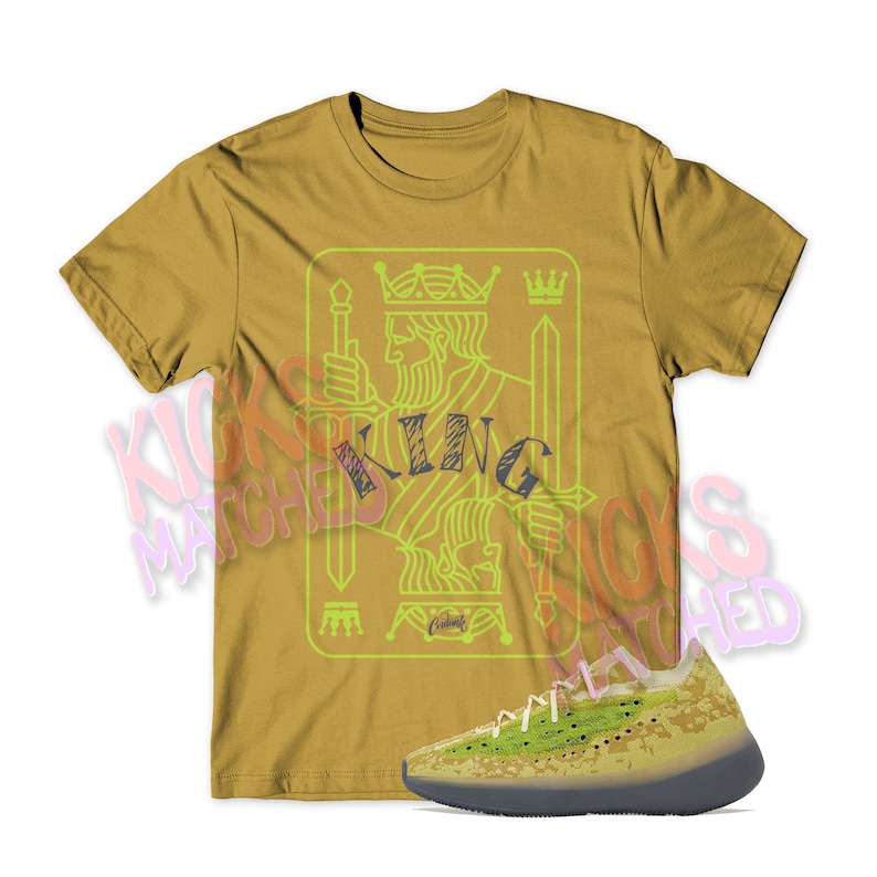 May include: A mustard yellow t-shirt with a green outline of a king of spades playing card graphic. The text "KING" is written in black inside the card graphic. A pair of green and yellow sneakers are shown below the t-shirt.