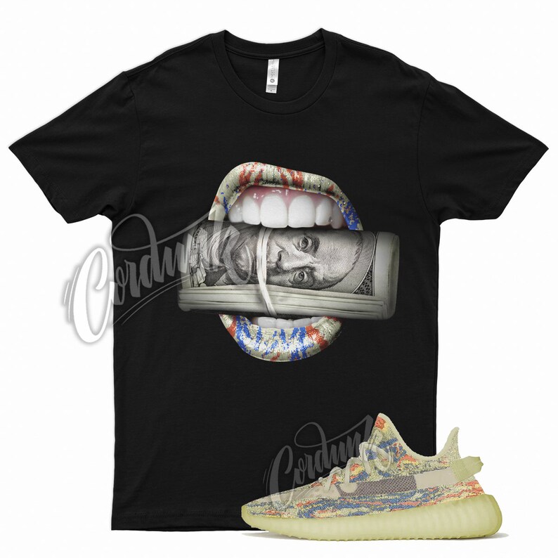 May include: Black t-shirt featuring a graphic of a mouth with red, white, and blue accents, holding a roll of hundred-dollar bills. A pair of multi-colored sneakers with a light yellow sole are in the foreground.