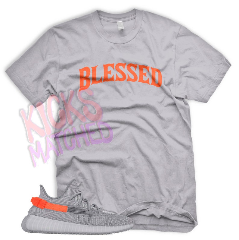 K&ouml;nnte beinhalten: Ein graues T-Shirt mit dem Wort "Blessed" in orangefarbenen Buchstaben. Unter dem Shirt befindet sich ein Paar graue und orangefarbene Sneaker.