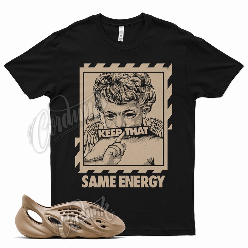 May include: Black t-shirt with a cherub graphic and the text "KEEP THAT SAME ENERGY." A pair of brown foam runners are in the foreground. The shirt has a crew neck and short sleeves.