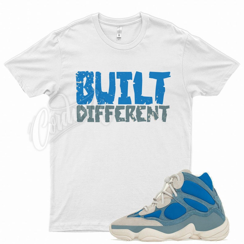 May include: A white t-shirt with the words "BUILT DIFFERENT" in blue and grey. A blue, grey, and cream-coloured trainer is in the foreground. The trainer has a chunky sole and a mix of suede and fabric.