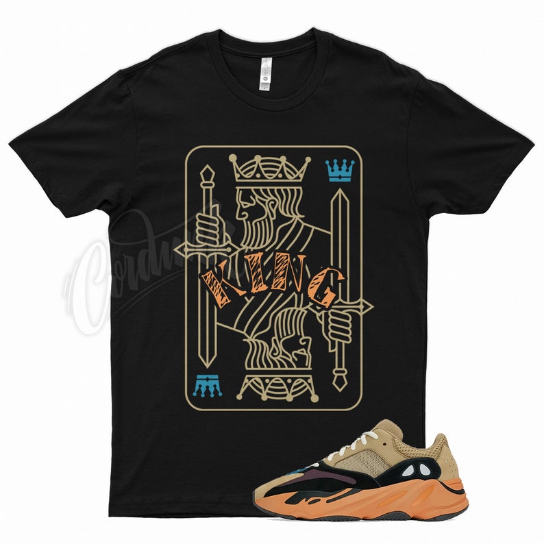 May include: Black t-shirt featuring a King card design with a crown and swords. The word "KING" is in orange. A tan, black, and orange sneaker is in the foreground.