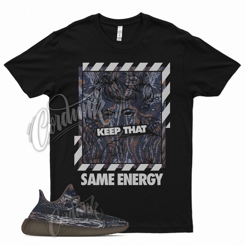 May include: Black crew neck t-shirt with a graphic design featuring a stylized face and the text "KEEP THAT SAME ENERGY" in white. A pair of matching sneakers with a brown sole are in the foreground.