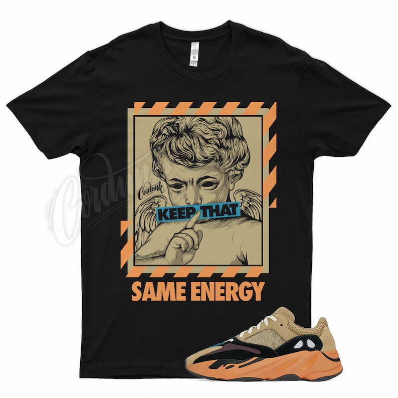 May include: Black t-shirt with an angel graphic, the text "KEEP THAT" on a blue banner, and "SAME ENERGY" in orange. A pair of tan, orange, and black trainers are in the foreground.