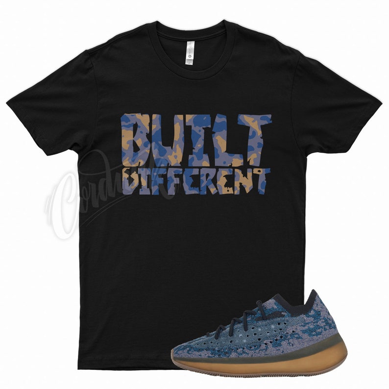 May include: Black t-shirt with the words "BUILT DIFFERENT" in a blue and tan camouflage pattern. A blue and tan camouflage sneaker with a brown sole is displayed below the shirt.
