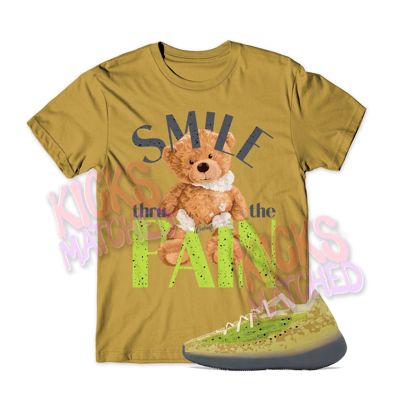 K&ouml;nnte beinhalten: Ein senfgelbes T-Shirt mit einem Aufdruck eines Teddyb&auml;ren, der ein Pflaster h&auml;lt, und dem Text "Smile thru the pain". Au&szlig;erdem ist ein Paar gr&uuml;n-gelber Sneaker mit einem gesprenkelten Design abgebildet.