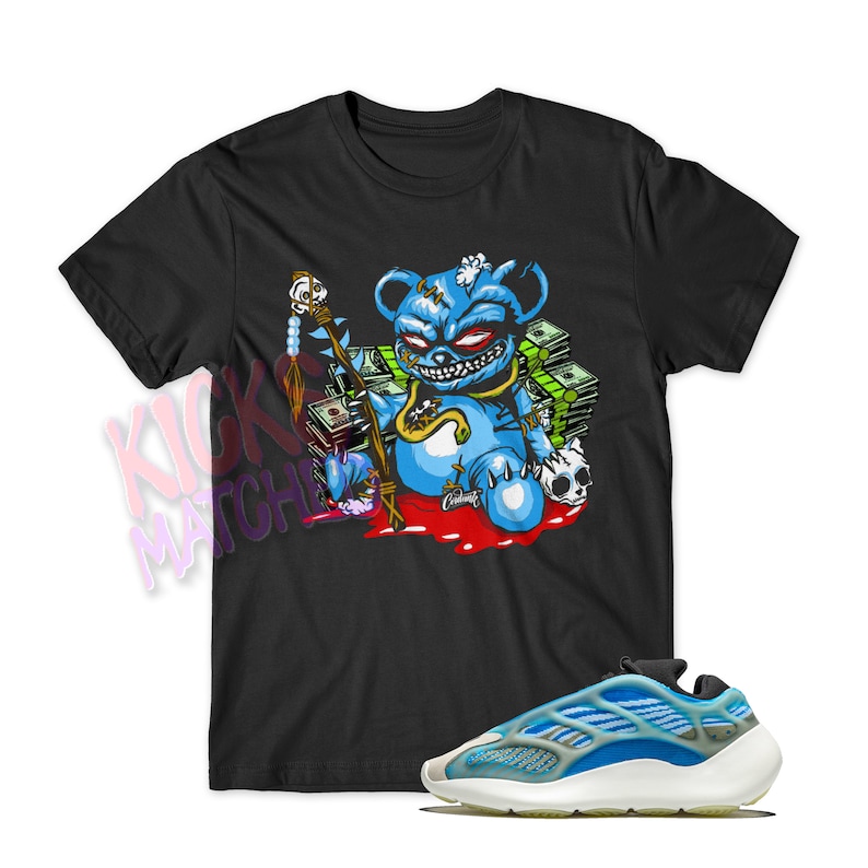 May include: Black t-shirt with a cartoon blue bear design, red eyes, gold chain, and staff, with money and skulls. A pair of blue and white trainers with a chunky sole are in the foreground. The text "Kicks Match" is visible.