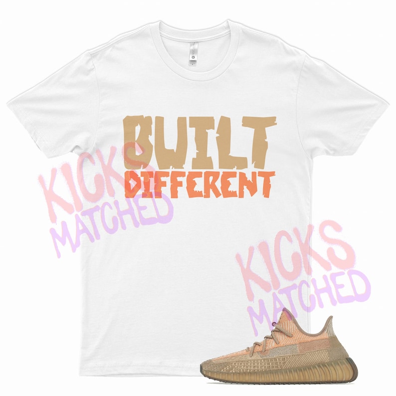 Pode incluir: Camiseta branca com o texto "BUILT DIFFERENT" em letras marrons e laranja. Um par de t&ecirc;nis Yeezy bege e marrom &eacute; mostrado abaixo da camiseta.