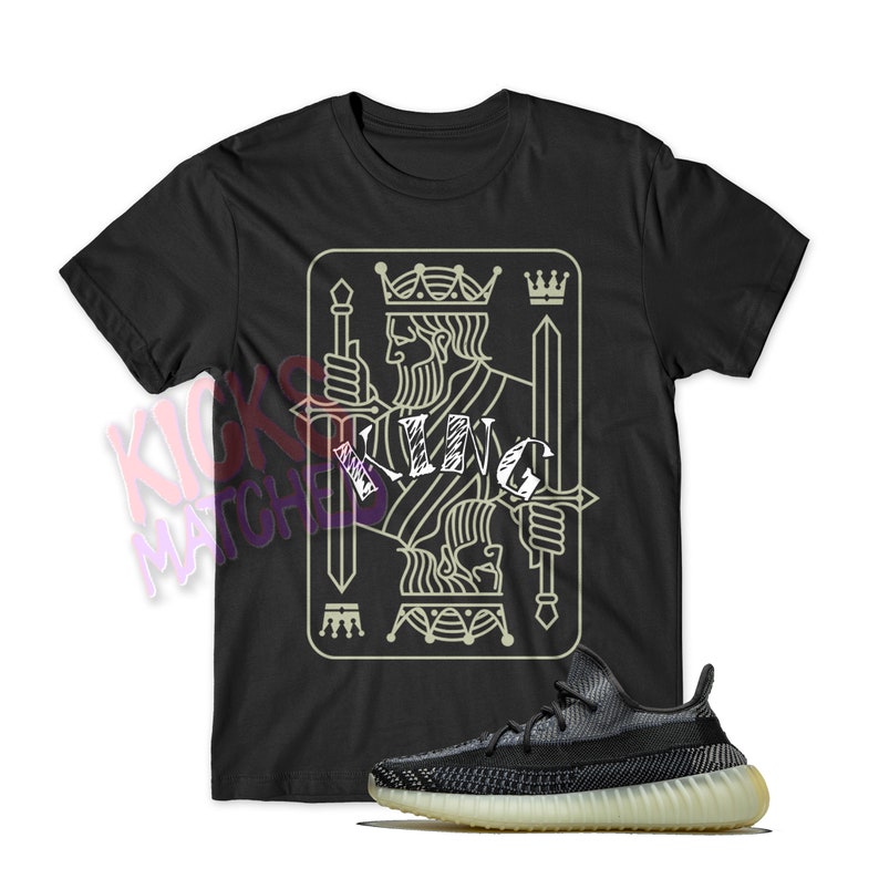 Puede incluir: Camiseta negra con un gr&aacute;fico de un rey de picas de la baraja de cartas con la palabra "KING" escrita en blanco. Un par de zapatillas negras y blancas se encuentran en la esquina inferior derecha de la imagen.