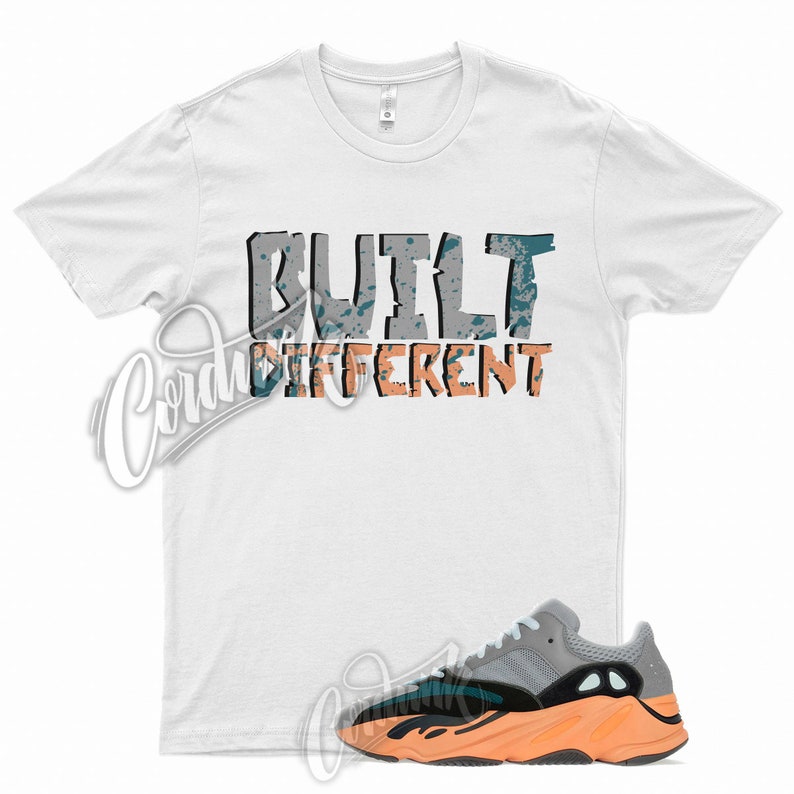 Op de afbeelding: Wit T-shirt met de tekst "BUILT DIFFERENT" in grijs en oranje met een turquoise splatter-effect. Een paar grijze, oranje en zwarte sneakers met een witte zool.