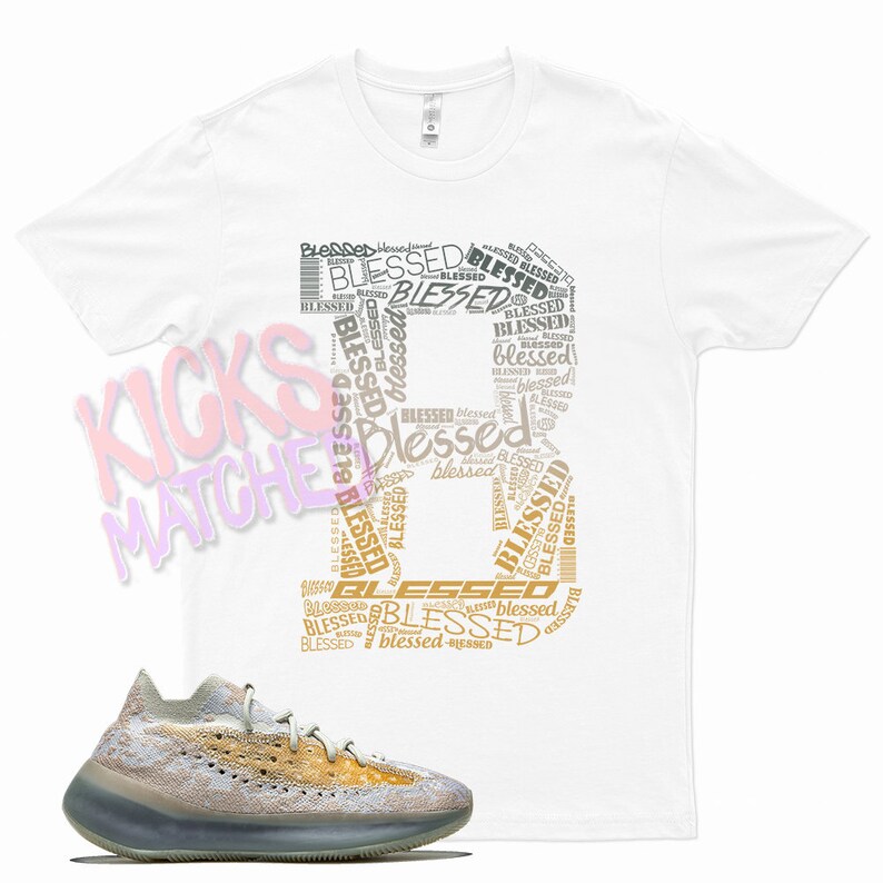 Op de afbeelding: Een wit T-shirt met een grafisch ontwerp van het woord "blessed" dat in verschillende formaten en lettertypen wordt herhaald. De grafiek is in tinten goud en bruin. Onder het T-shirt is een paar lichtgrijze en beige sneakers met een lichtblauwe zool te zien.