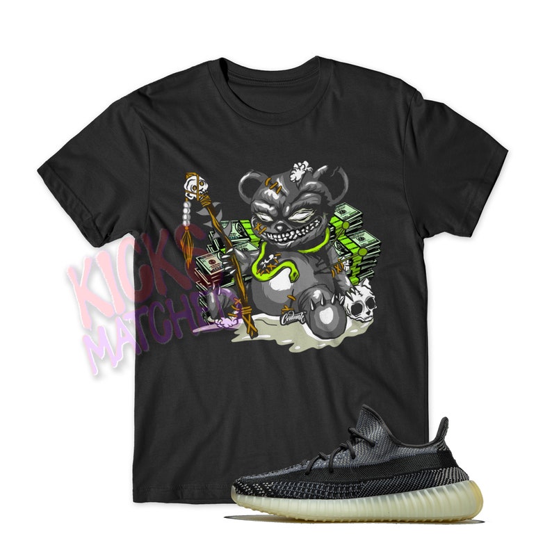 May include: Black t-shirt featuring a cartoon illustration of a green and black bear wearing a necklace and holding a staff. The bear is sitting on a pile of money. The t-shirt is paired with black and white trainers with a green stripe.