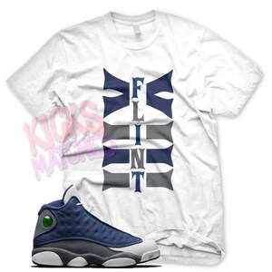 May include: White t-shirt with the word "FLINT" printed in blue and gray. A pair of blue and gray Air Jordan 13 sneakers.