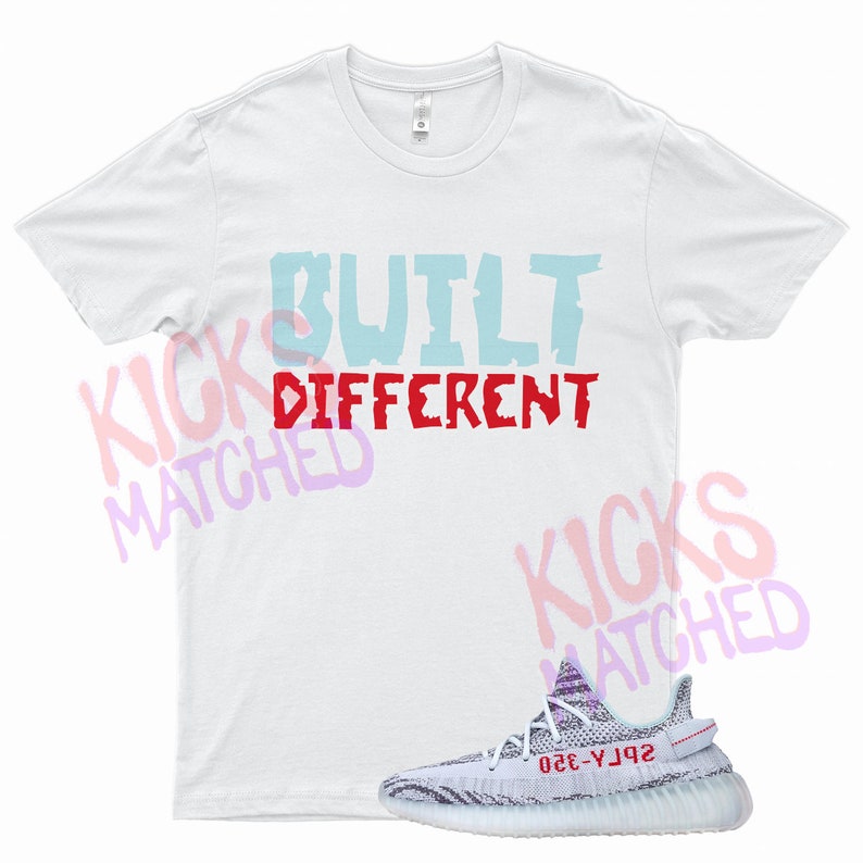Puede incluir: Camiseta blanca con el texto "BUILT DIFFERENT" en letras azules y rojas. Un par de zapatillas blancas y grises con una franja roja y detalles azules.