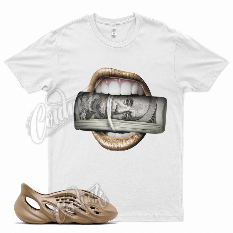May include: White t-shirt featuring a graphic of gold lips holding a roll of $100 bills. A pair of brown foam runners are in the foreground. The shirt has a crew neck and short sleeves.