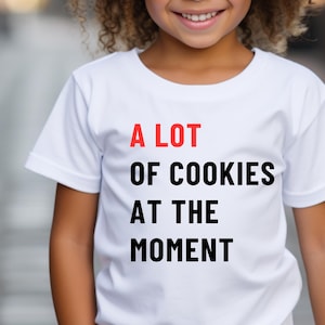 May include: A young girl wearing a white t-shirt with the text "A LOT OF COOKIES AT THE MOMENT" printed in black and red.