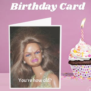 May include: A birthday card with the text "Birthday Card" at the top. The card features a doll with a displeased expression and the text "You're how old?" A cupcake with white frosting, sprinkles, and a lit candle is to the right.