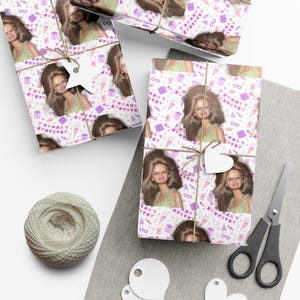 May include: Gift-wrapped presents with a white and pink patterned paper featuring a doll image, party flags, and gift boxes. Each package is tied with twine and a white tag. A ball of twine, scissors, and blank tags are also present.
