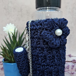 May include: A navy blue crochet water bottle holder with a flower accent and pearl center. The holder has a silver chain strap and a matching small pouch. A clear water bottle with a black lid is inside the holder.