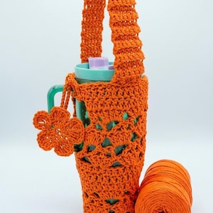 May include: A bright orange crocheted water bottle holder with a long handle and a matching flower-shaped charm attached to the side. The holder features an openwork pattern and contains a teal plastic water bottle with a light purple lid.