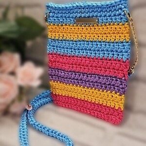 May include: A colorful crocheted crossbody bag with a blue strap and a gold chain. The bag has stripes of blue, yellow, pink, and purple.