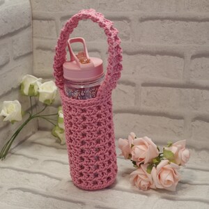 May include: A pink crocheted water bottle holder with a matching handle. The holder encases a clear water bottle with a pink lid. The bottle is filled with a floral pattern. Artificial pink roses and white roses are in the background.