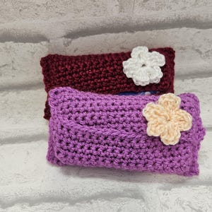 Puede incluir: Dos estuches de ganchillo hechos a mano. El estuche superior es de color burdeos con una flor blanca. El estuche inferior es morado con una flor color crema. Ambos estuches son rectangulares y tienen un patrón de puntada texturizada.