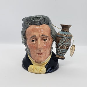 May include: A ceramic mug shaped like a man's head with a small blue and brown vase attached to the side. The mug is decorated with a white collar and a yellow bow tie. The vase has a gold trim and intricate designs. The mug has a label that reads "Royal Doulton".