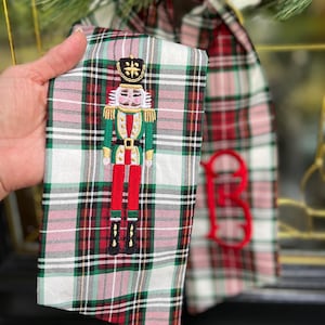 May include: A Dress White Stewart plaid fabric stocking featuring a 6-inch embroidered nutcracker figure in green, red, and gold thread, alongside a large red embroidered fishtail monogram letter B on the adjacent fabric section.