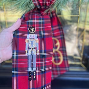 May include: A red and green plaid fabric ribbon with a gray embroidered nutcracker design. The ribbon is tied in a bow and is hanging from a Christmas wreath.