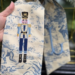 May include: A beige fabric ribbon featuring a blue toile de jouy pattern and an embroidered nutcracker figure. The nutcracker wears a blue and gold uniform with a black hat. A blue embroidered letter L is visible on the adjacent fabric section.
