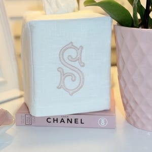 May include: A square white linen tissue box cover featuring a centered embroidered letter S in powder pink thread. The cover is placed on top of a pink book with the text COCO CHANEL printed on the spine in black capital letters.