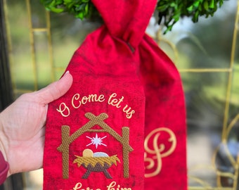 May include: A red fabric door hanger featuring a mottled texture and embroidered gold text that reads O Come let us adore him. The design includes a brown manger, a white star, and a gold monogram letter. It hangs from a green wreath.