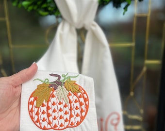 May include: A white fabric towel featuring an embroidered orange pumpkin with a textured pattern and green leaves. The pumpkin design is stitched in orange thread with a repeating triangular motif. A monogram is visible on the lower section.