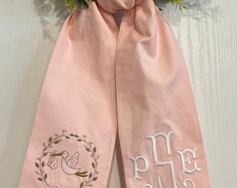 May include: A pink fabric sash with white embroidery. One side features a stork carrying a baby in a basket, surrounded by a wreath of leaves. The other side has the monogram "PJE".