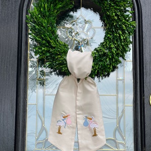May include: A white fabric door wreath with two embroidered storks carrying baby bundles, one blue and one pink. The wreath is hung on a black door with a glass window.