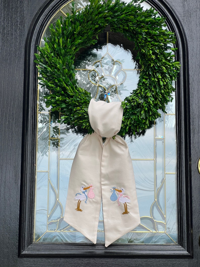 May include: A white fabric door wreath with two embroidered storks carrying baby bundles, one blue and one pink. The wreath is hung on a black door with a glass window.