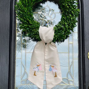 May include: A white fabric door swag with two embroidered storks carrying baby bundles, one pink and one blue. The swag is tied around a green boxwood wreath.