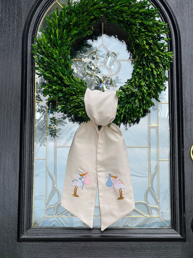 May include: A white fabric door swag with two embroidered storks carrying baby bundles, one pink and one blue. The swag is tied around a green boxwood wreath.