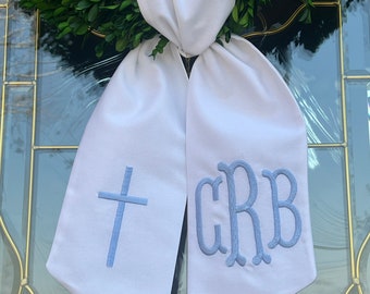 May include: A white fabric sash draped over a green boxwood wreath. The left side of the sash features an embroidered light blue cross, while the right side displays an embroidered light blue monogram with the letters CRB in a script font.