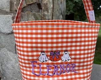 May include: A fabric bucket with an orange and white gingham pattern and a matching handle. The front features an embroidered design of two white ghosts flanking an orange pumpkin with a purple bow, and the name Eleanor in purple text.