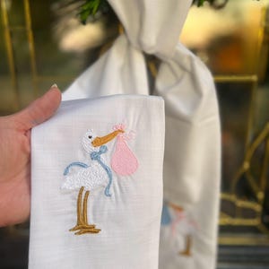 May include: White linen-look fabric with a white stork embroidered on it carrying a pink bundle. The stork has blue ribbon accents. The fabric is folded in half and held in a hand.