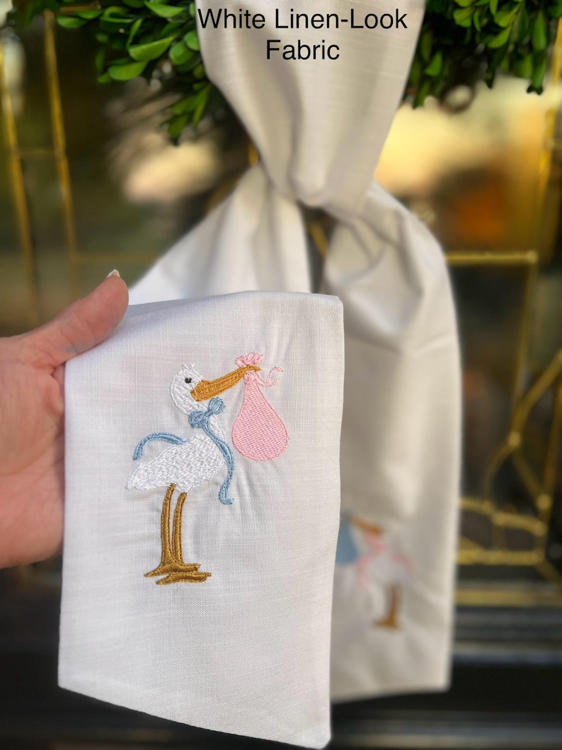 May include: White linen-look fabric with a white stork embroidered on it carrying a pink bundle. The stork has blue ribbon accents. The fabric is folded in half and held in a hand.