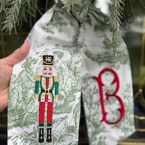 May include: A white fabric sash featuring a green toile pattern, an embroidered nutcracker figure, and a large red monogram letter B. The sash includes text that reads Fern Green Toile 52" Sash Red Monogram, indicating a length of 52 inches.