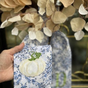 May include: A rectangular fabric piece featuring a white embroidered pumpkin with a green stem and leaves, set against a blue and white damask pattern. The text "Elegant White Pumpkin on Blue Damask Fabric" is visible at the top of the image.