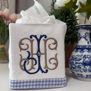 May include: A white linen tissue box cover with a blue and white gingham trim. The cover features a brown and blue embroidered monogram with the letter "H".