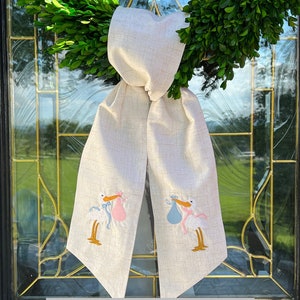 May include: A creamy beige textured fabric door wreath with a bow and two embroidered storks carrying baby bundles, one pink and one blue.
