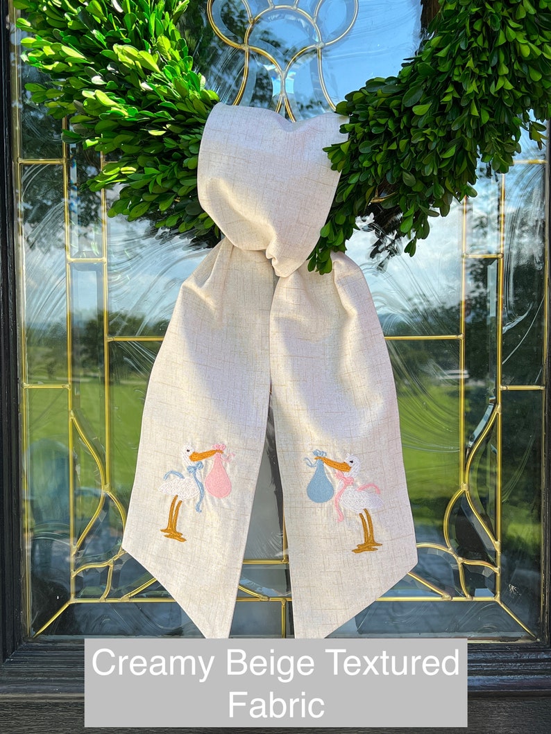 May include: A creamy beige textured fabric door wreath with a bow and two embroidered storks carrying baby bundles, one pink and one blue.