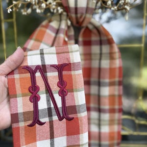 May include: A rectangular fabric piece featuring an autumn plaid pattern in shades of orange, cream, brown, and green. The fabric is embroidered with a large burgundy letter N. Text on the image reads: Autumn Plaid Fabric, Burgundy Letter, Cranberry Knot.